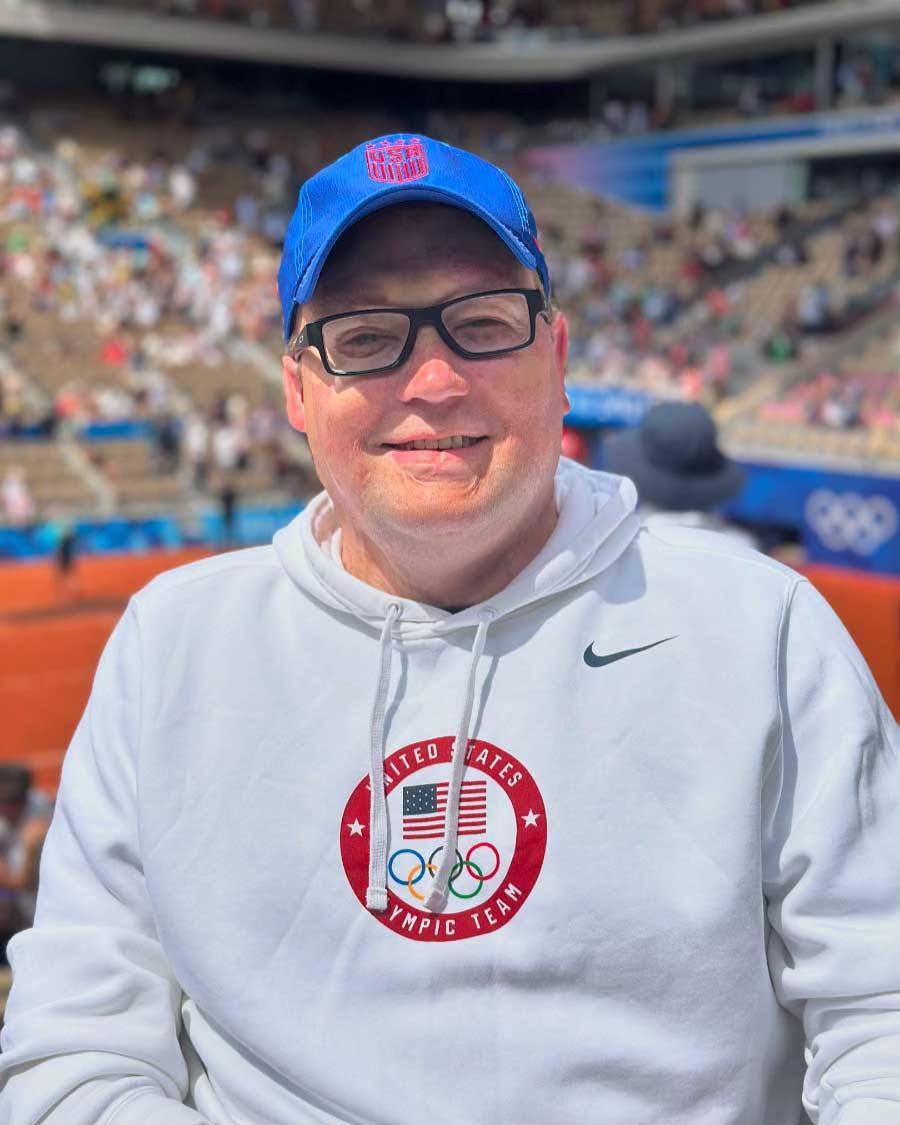 John wearing a white United States Olympic team hoodie with a blue USA baseball cap at a tennis stadium.