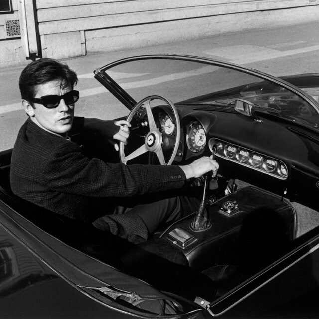 A black and white photo of Alain Delon driving a convertible sports car in sunglasses, looking at the camera with one hand on the steering wheel and the other on the gearshift.