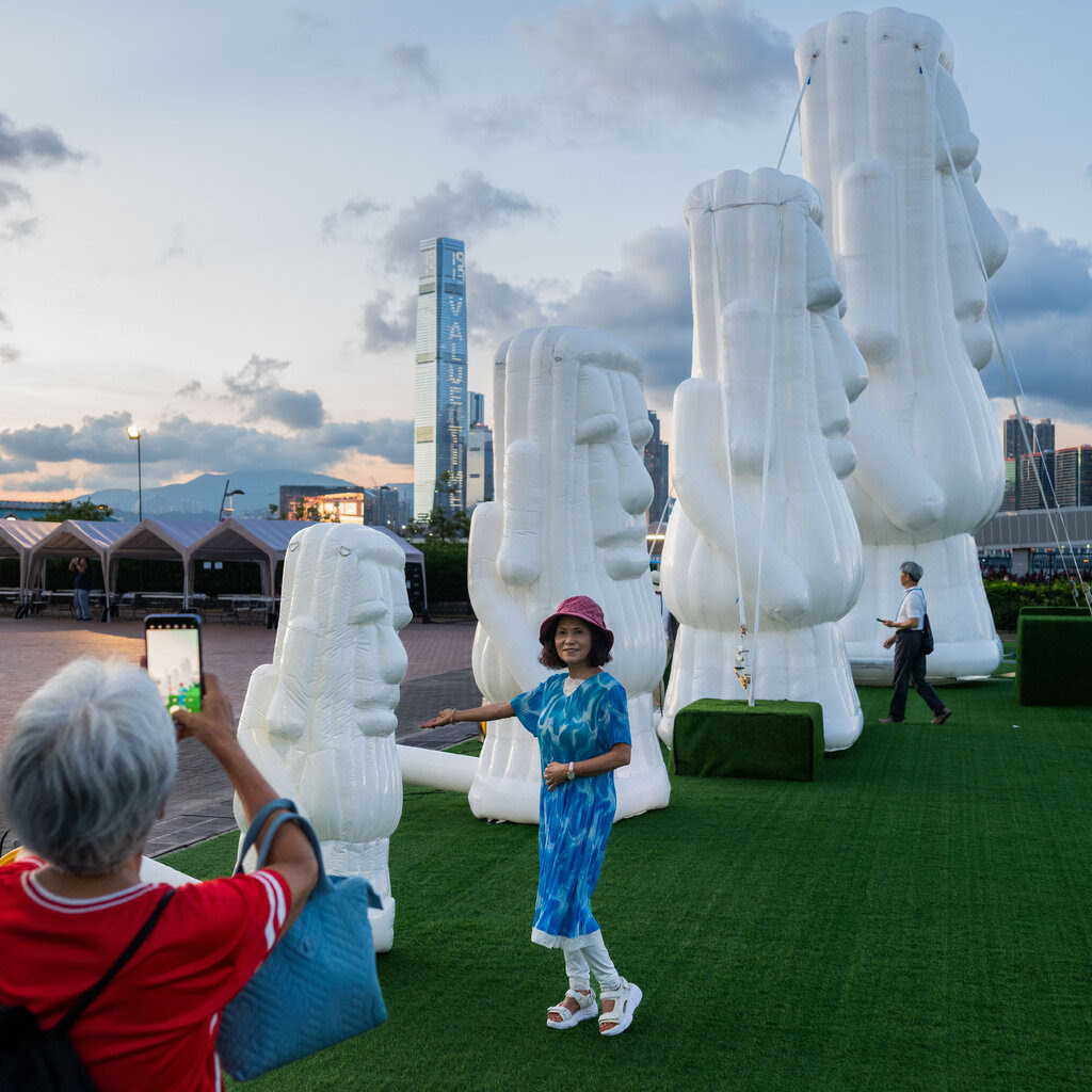 A woman stands in front of one of a series of an inflatable white replicas of elongated heads as someone photographs her on a phone.