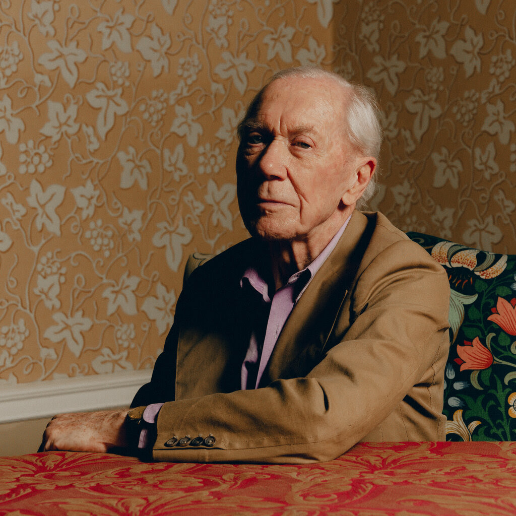 A man wearing a brown coat and a purple shirt sits in front of wallpaper patterned with grapevines.