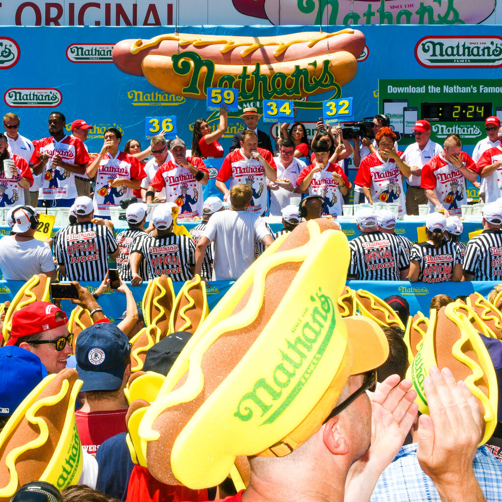 A large crowd of spectators, many wearing hot dog-shaped hats, watch as a lineup of competitive eaters consumes hot dogs. 