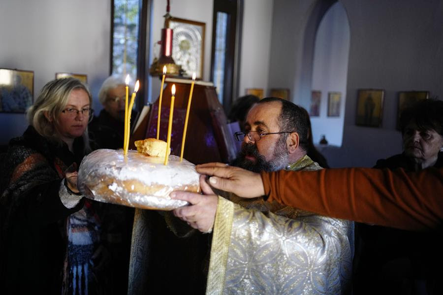 A man in religious robes holds a loaf of bread with candles in it. There are people gathered around him laying their hands on the loaf as well.