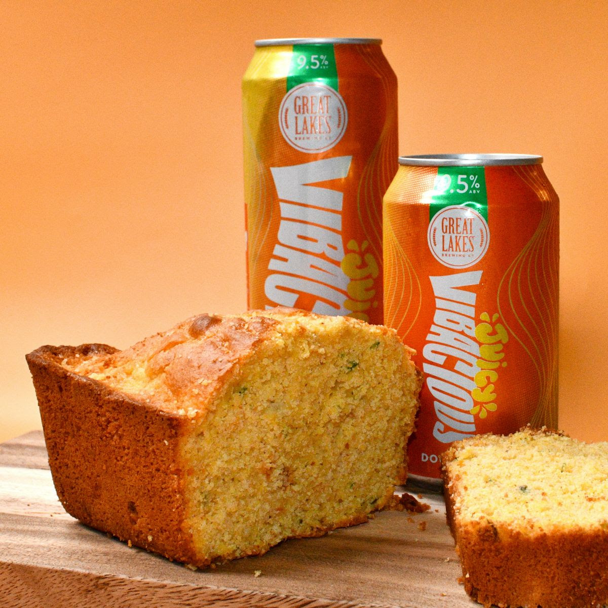 Peach Jalapeno Cornbread made with Juicy Vibacious Double IPA. 12 oz. and 19.2 oz. cans of the beer next to the cornbread. Wood surface, orange background.