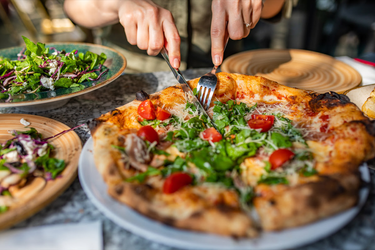 A pair of hands slicing a pizza with a fork and knife.