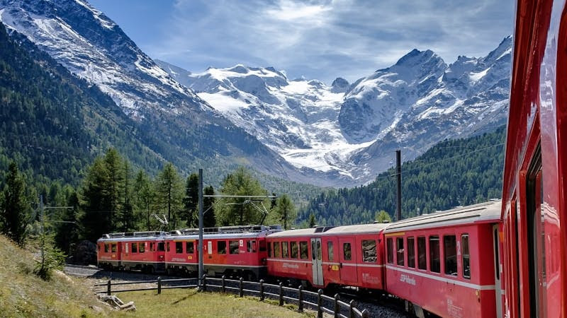 red and white train near green field viewing mountain and green trees