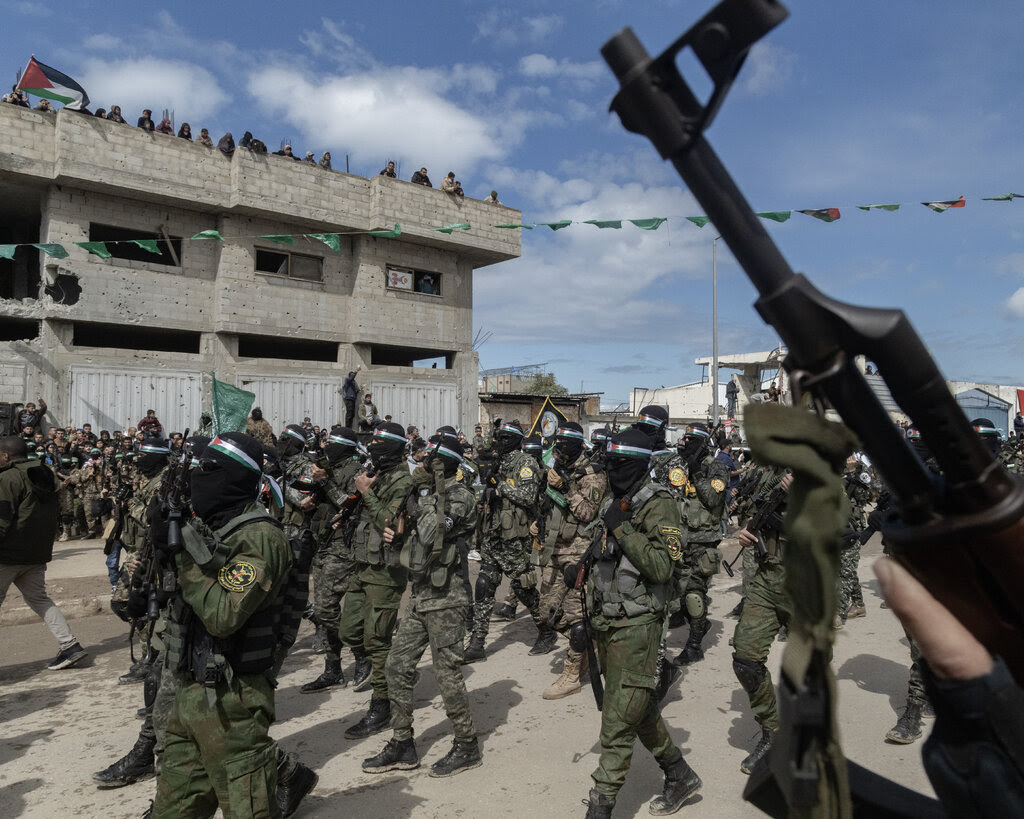 Armed fighters in uniform marching past a concrete building.