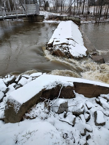 water rushes through a broken part of a snow-covered cement dam on a dark green river