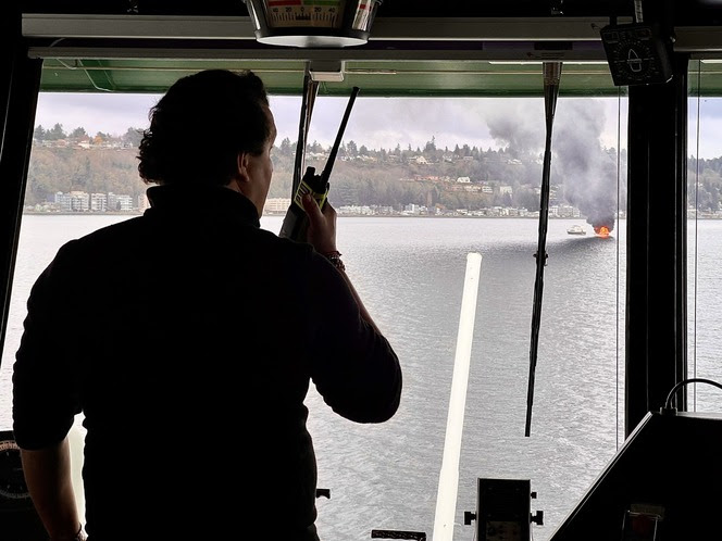 Person in ferry wheelhouse observing a burning boat with smoke on the water and shoreline visible in the background