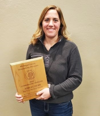 a woman with medium length dark blond hair and in a gray, long-sleeved shirt smiles as she holds a rectangular, engraved plaque