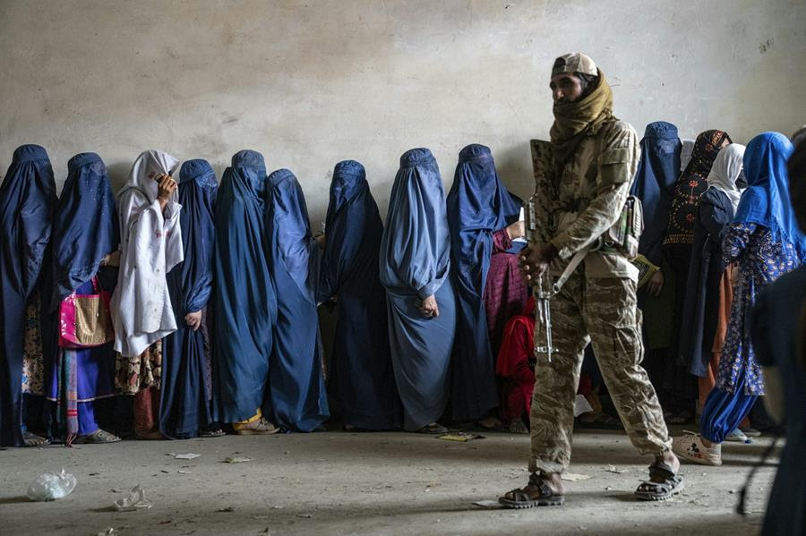 A man with a gun stand next to a long line of women who are covered from head to toe in a hijab.