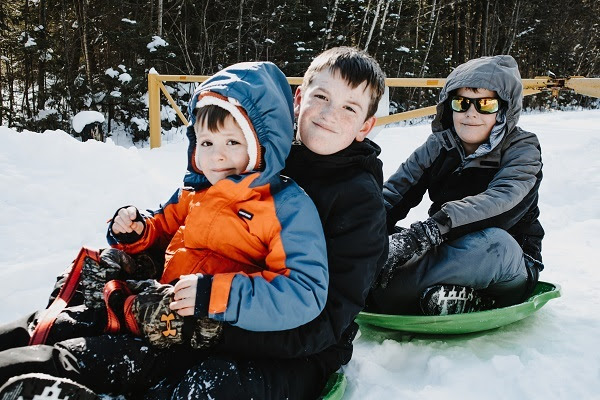 three young boys in winter coats and snowpants sit on green saucer sleds on the snow-covered, forested ground