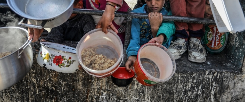 Palestinian children in Gaza wait behind railings with their arms outstretched holding empty containers for food.