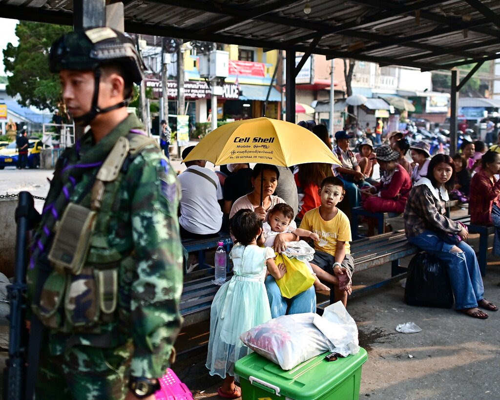 A woman with three children sits on a bench and holds up a yellow umbrella. A soldier stands near her.