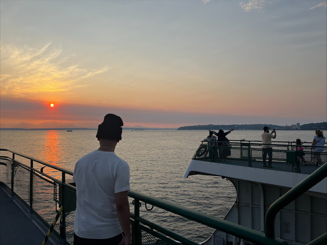 Person in a beanie looking out on the outdoor deck of a ferry at the setting sun with a few other people also on the deck in the distance