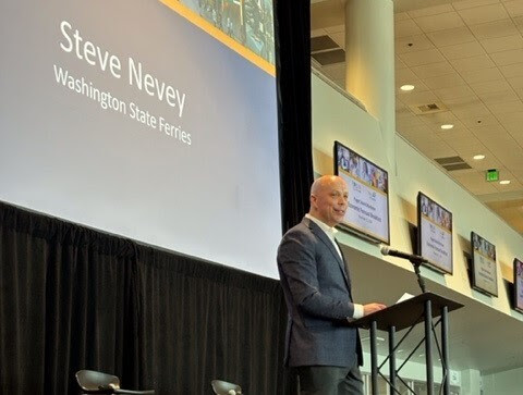 A man speaks at a podium in a large room with a screen displaying "Steve Nevey, Washington State Ferries"