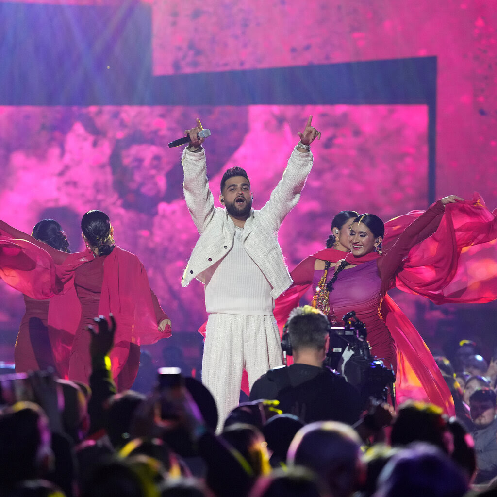 A man with a microphone in an all-white outfit holds his hands out, surrounded by dancers in pink and purple against a pink and purple backdrop.