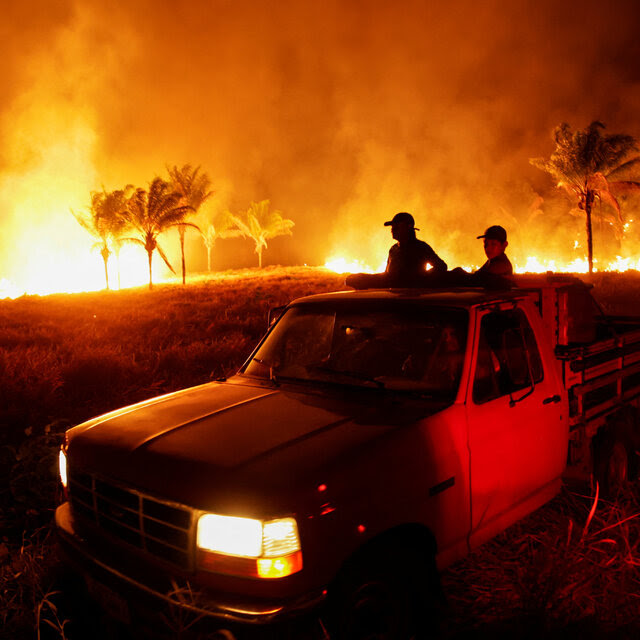 Two people in the back of a pickup truck look out over a fire at night in a rural area.