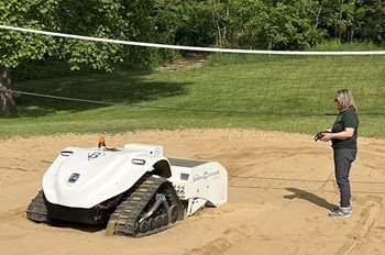 a woman in dark green shirt and pants stands next to a low, white vehicle with tracked runners. They're on a sand volleyball court