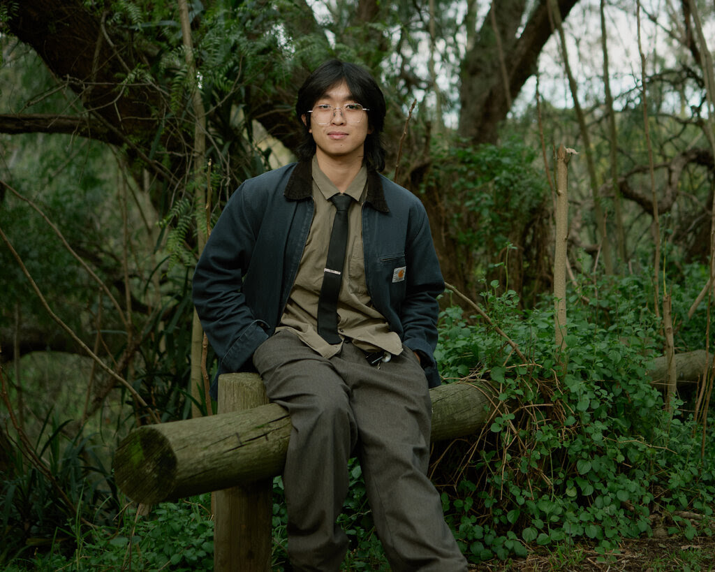 A man with wire-framed glasses sits on a log in a wooded area with his hands in the pockets of a blue work jacket.