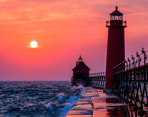 A brilliant orange sunset casts a lighthouse at the end of a pier in hues of pink, blue and purple.
