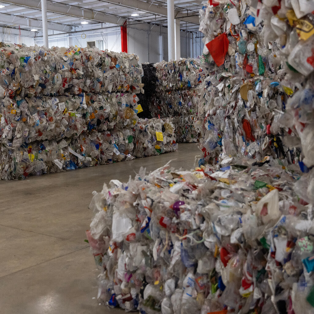 Bales of crushed plastic are piled in neat rows on a concrete floor inside a white-walled warehouse.