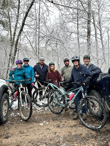 A small group of men and women in winter outdoor gear and helmets stand with mountain bikes on a snowy, forested trail