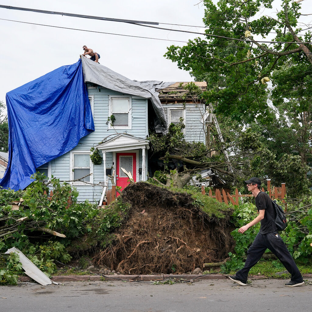 Uprooted trees in front of a house with tarps over its roof.