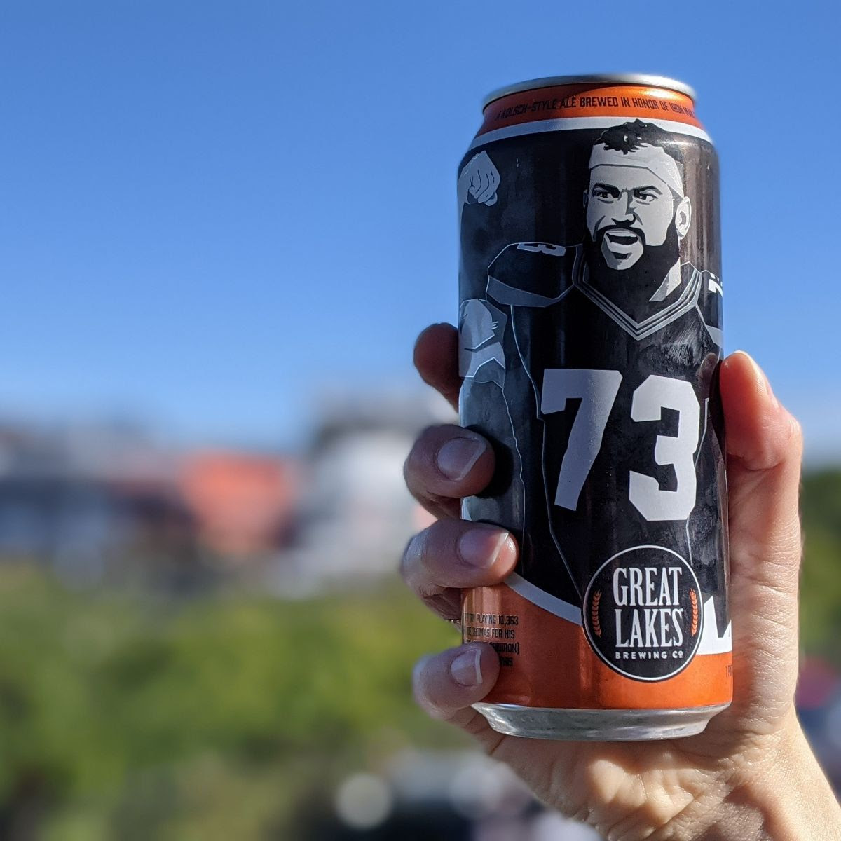 Close up of person holding a 16 oz. Can of 73 Kolsch in front of a downtown Cleveland backdrop.