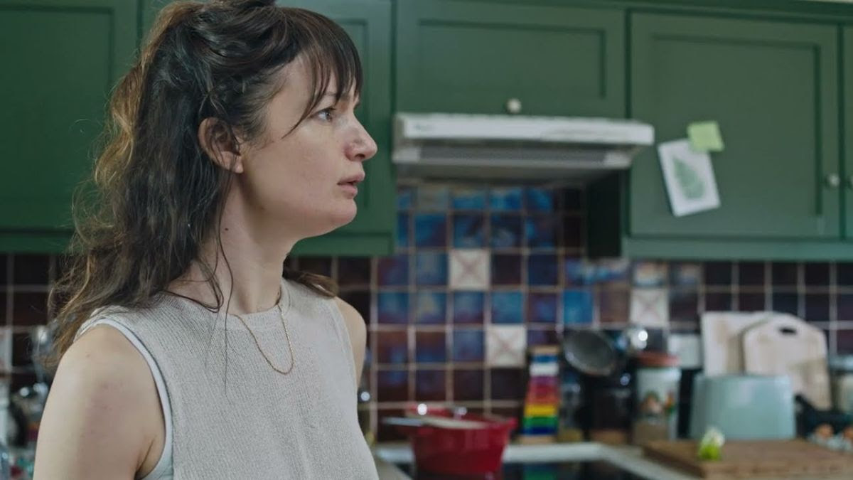 A woman with brown hair and wearing a grey sleeveless top stands left of centre, facing right with a distinct look of apprehension. In the background is a kitchen.