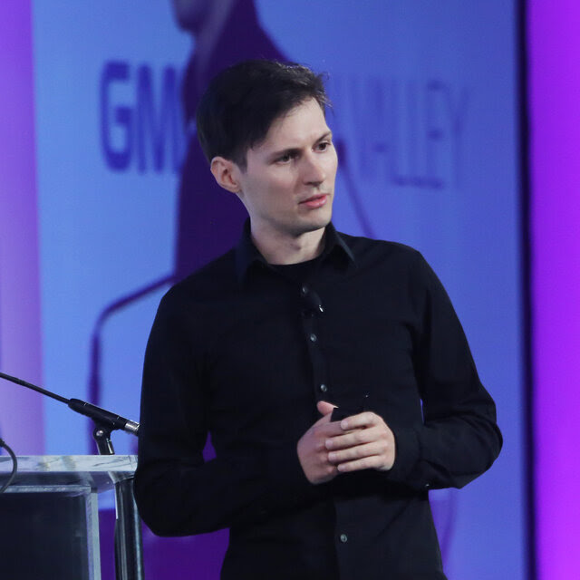 Pavel Durov, wearing a dark button down shirt and a dark t- shirt, standing in front a lectern. 