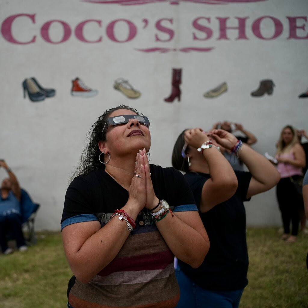 A woman, wearing eclipse glasses, looks up at the sky with her hands clasped together in prayer. 