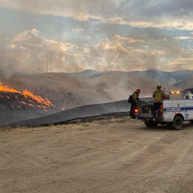 Two men in the back of a fire department pickup truck are parked near scorched earth and a patch of ground on fire.