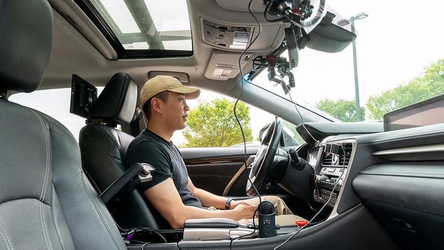 A student sits in a vehicle’s driver’s seat, hands in lap and surrounded by several gadgets hooked up to the vehicle’s interior