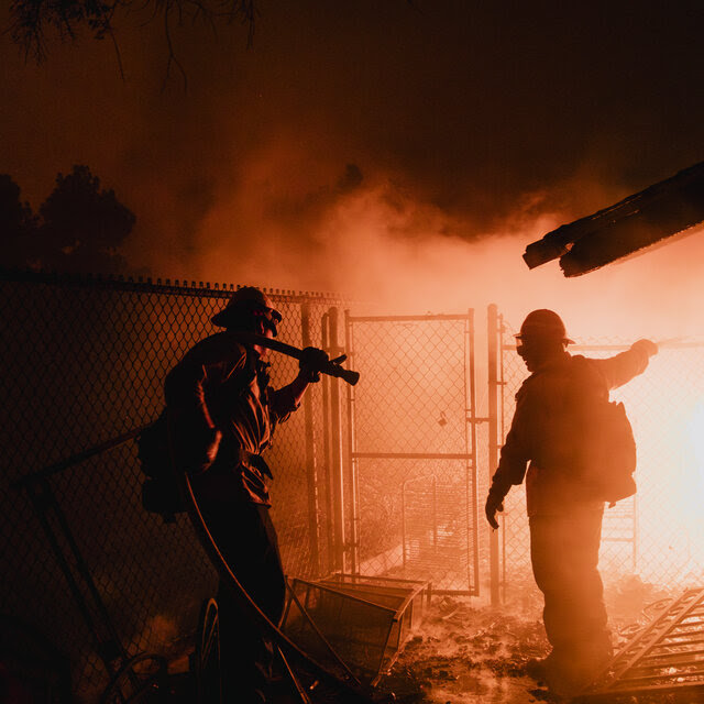 Two firefighters stand next to a fenced area, as an orange glow radiates from a fire.