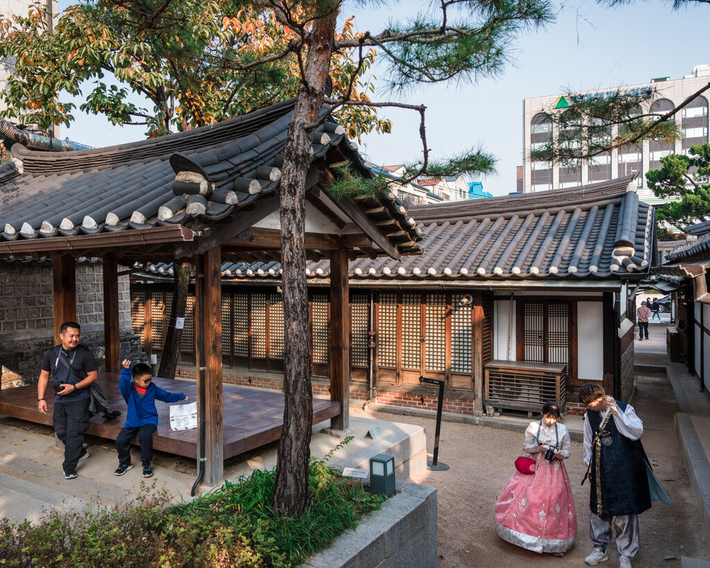 Four people, two wearing modern clothing, two in traditional Korean attire, walk in the shady courtyard of a hanok.