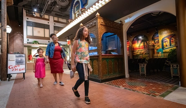 a woman and two young girls in summer clothing walk down a brick pathway in a museum, lined with ornate exhibits and bulb lights