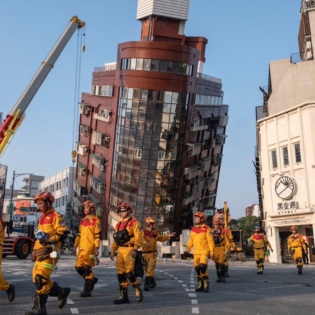 People in yellow suits and red helmets approach a building that is leaning toward the street. 