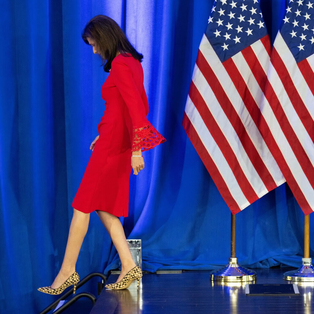 Nikki Haley in a red dress and yellow and black high heels walks off a stage. On the stage are large Americans flags.