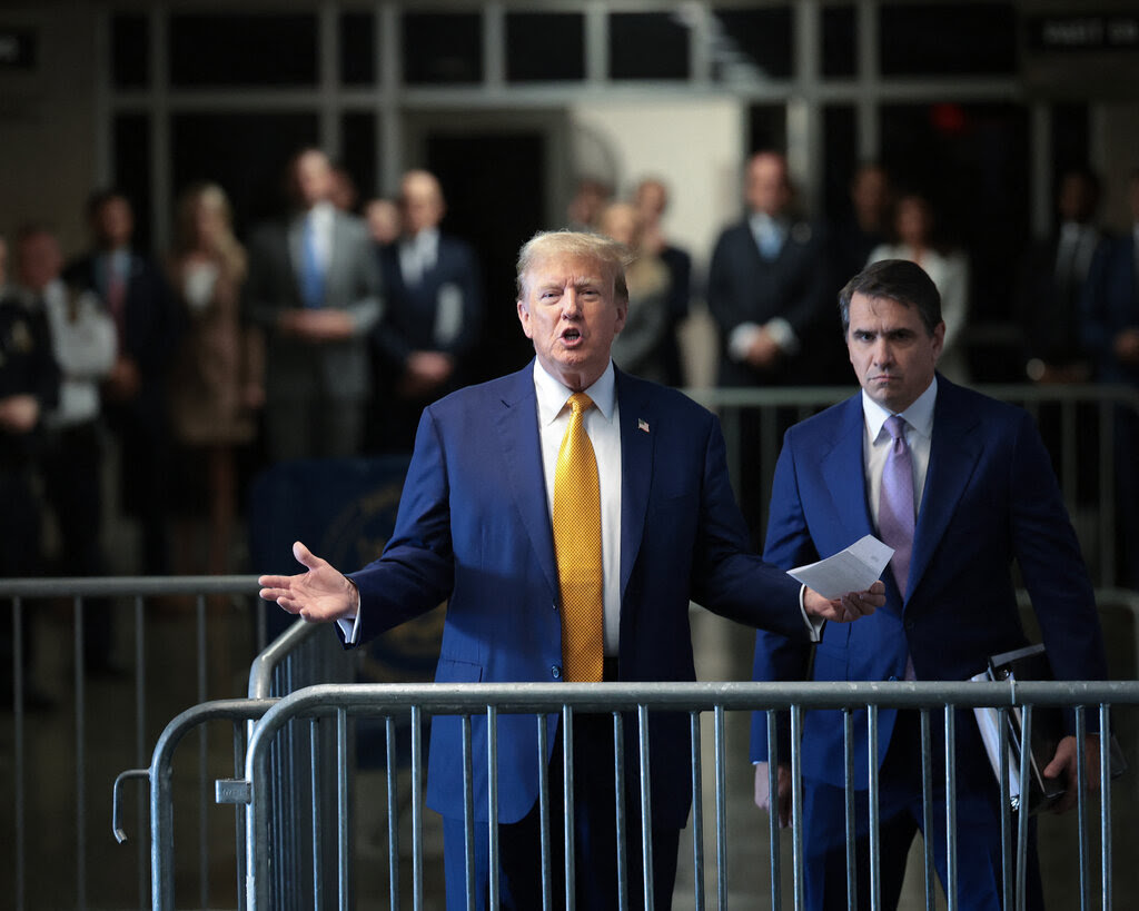 Donald Trump and Todd Blanche, both in blue suits, standing in front of barricades. Trump is gesturing with both hands and speaking.