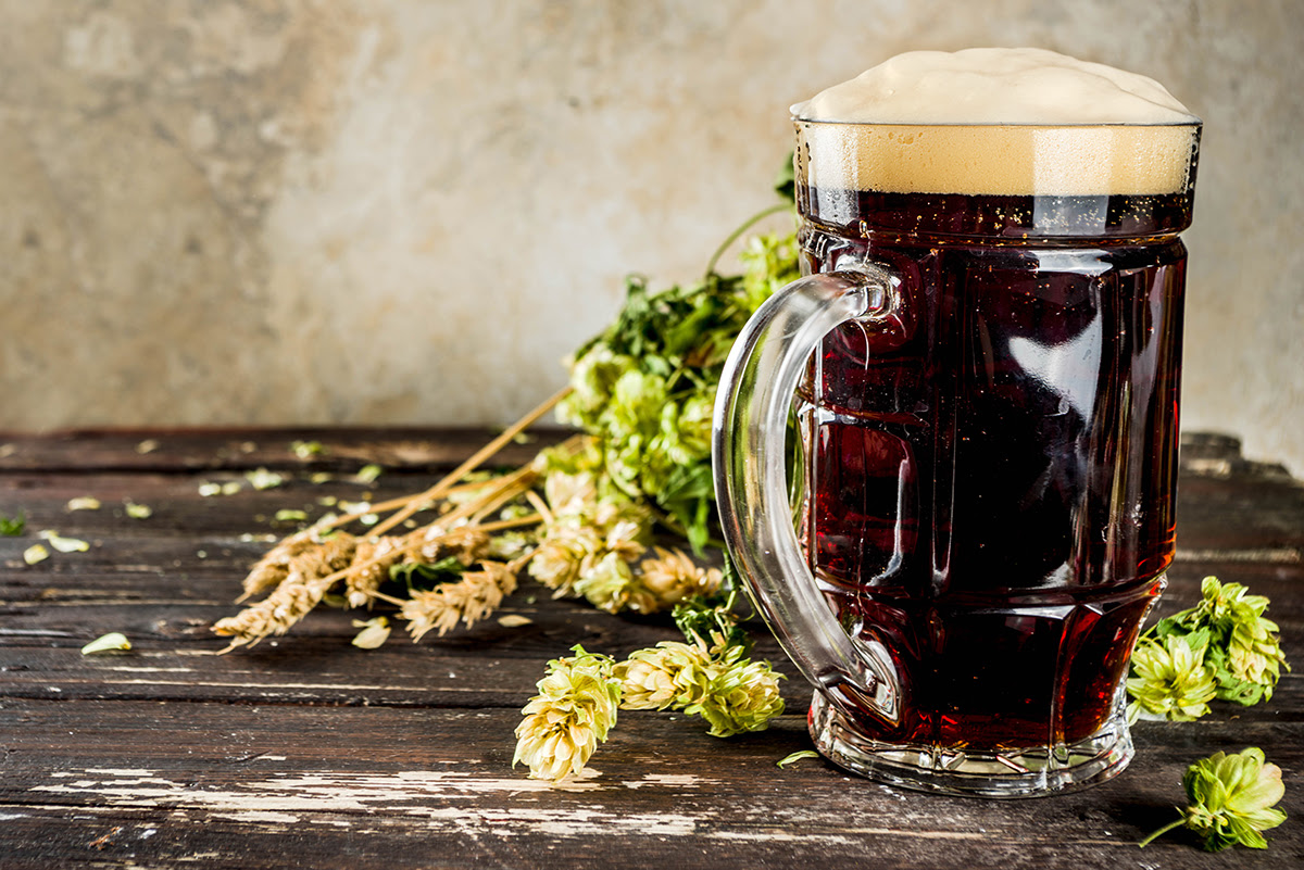 A glass of dark beer on a rustic table with hops and wheat.