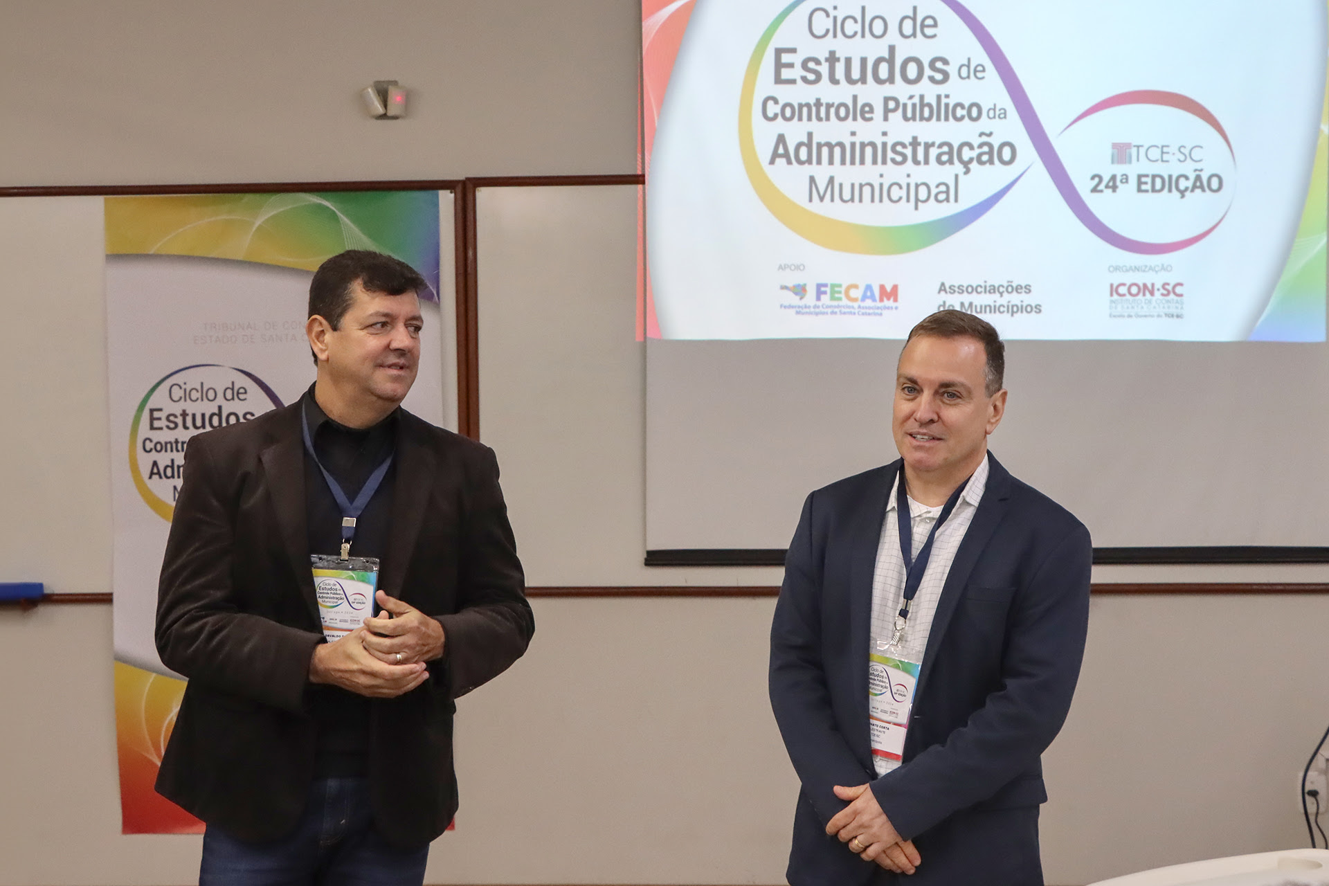 Foto de 2 servidores do TCE. Osvaldo Faria de Oliveria - a esquerda. Renato Costa, a direita. Atrás deles uma projeção de banner com a logomarca do Ciclo de Estudos.