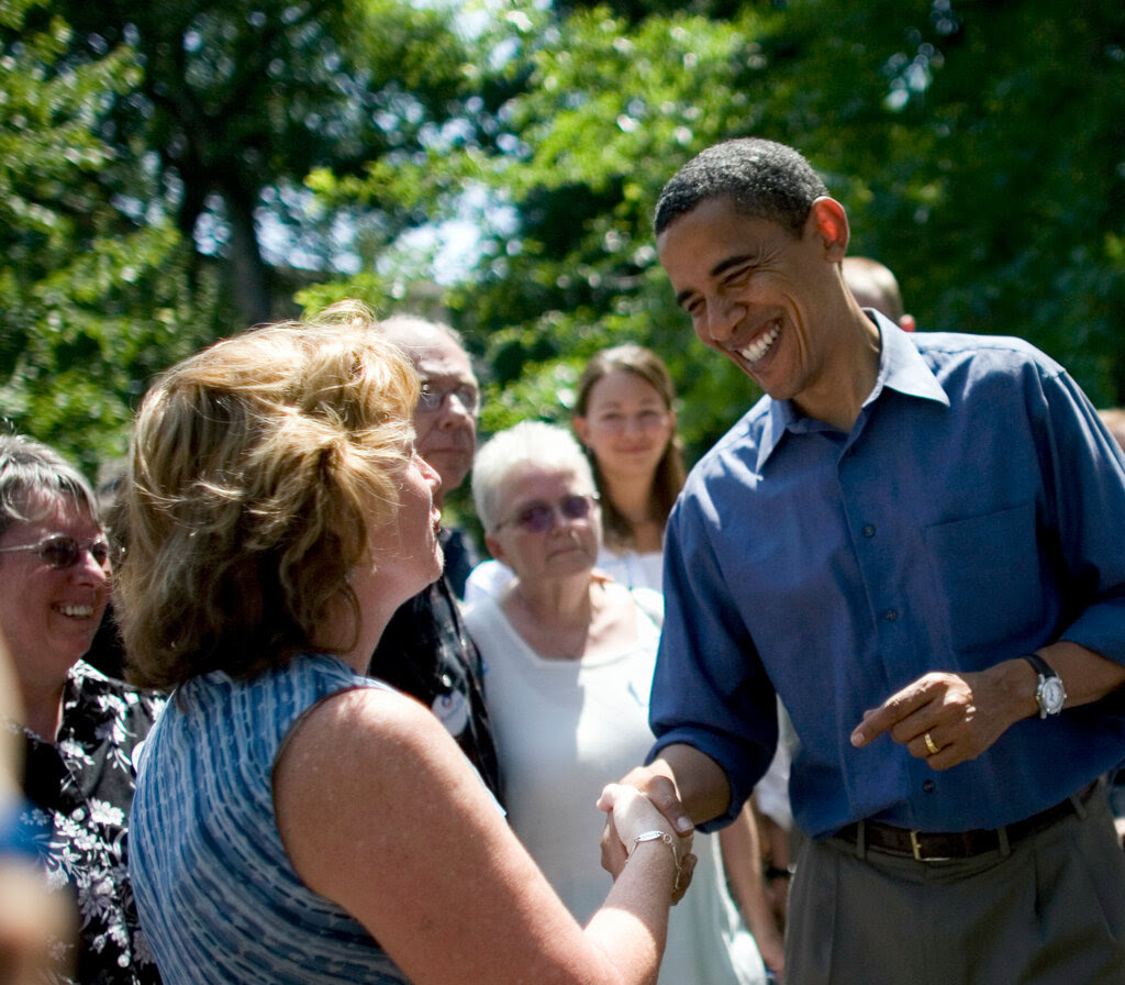 Barack Obama is smiling and shaking hands with someone as a small crowd looks on.