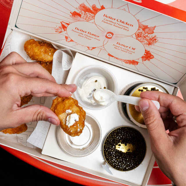 A pair of hands hovers over a box containing chicken nuggets and caviar, with a spoonful of sour cream.  