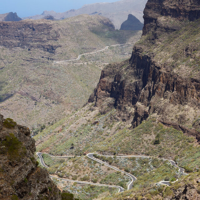 Winding roadways are visible in a large, steep ravine. A few people stand near the edge of a bluff in the foreground, surveying the landscape. 