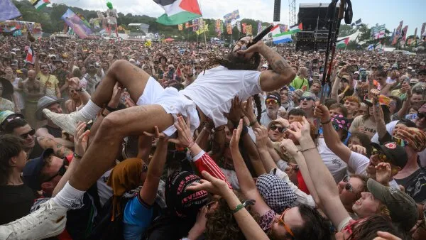 Bobby Vylan of Bob Vylan crowdsurfs in front of the West Holts stage during day four of the Glastonbury festival 2025 on June 28 in Glastonbury, England. Vylan can be seen wearing a white-colored short-sleeve shirt, shorts, socks and sneakers. In the crowd, several Palestinian flags can be seen waving.