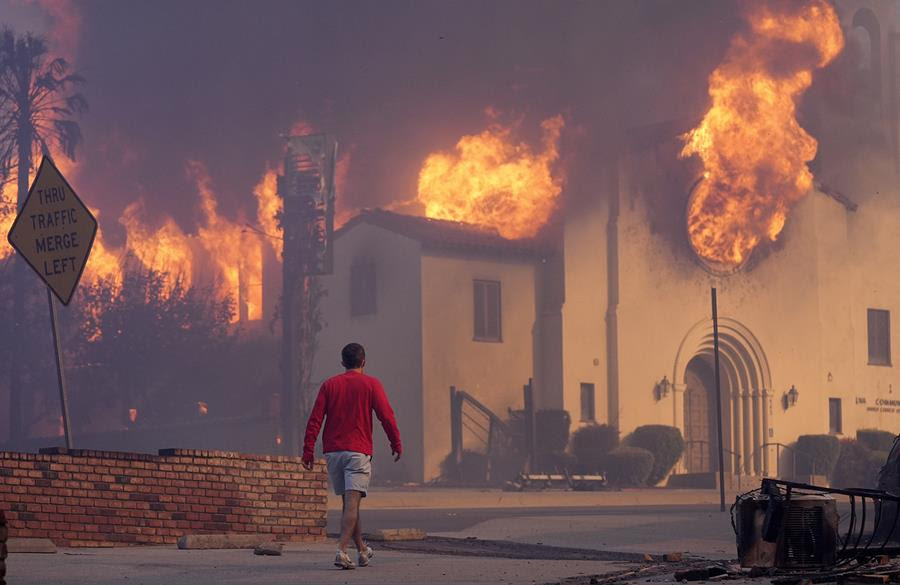 A man walks down a street littered with debris and ash. The Altadena Community Church is burning in the background with smoke and fire coming out of its windows.