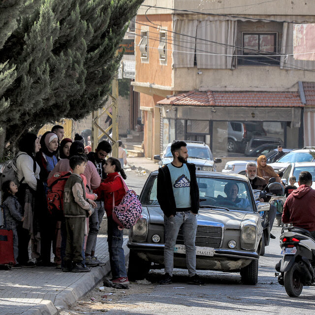 A crowd of people carrying bags stands on a sidewalk.