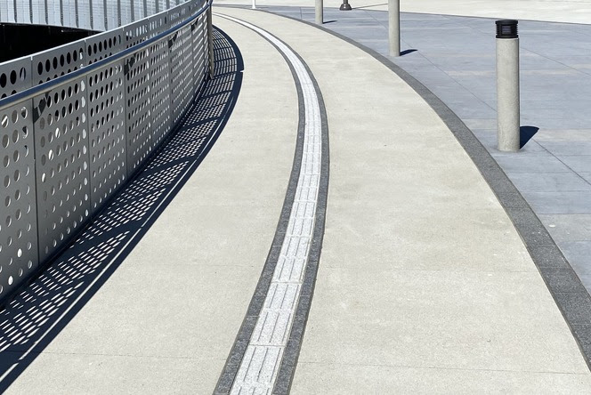 White directional tactile ground surface indicators along a walkway between a railing and short light pillars