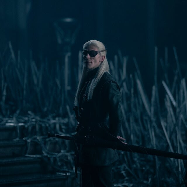 A young man with long blond hair, wearing an eye patch, stands in a dark room filled with swords, looking toward a blurry figure in the foreground.