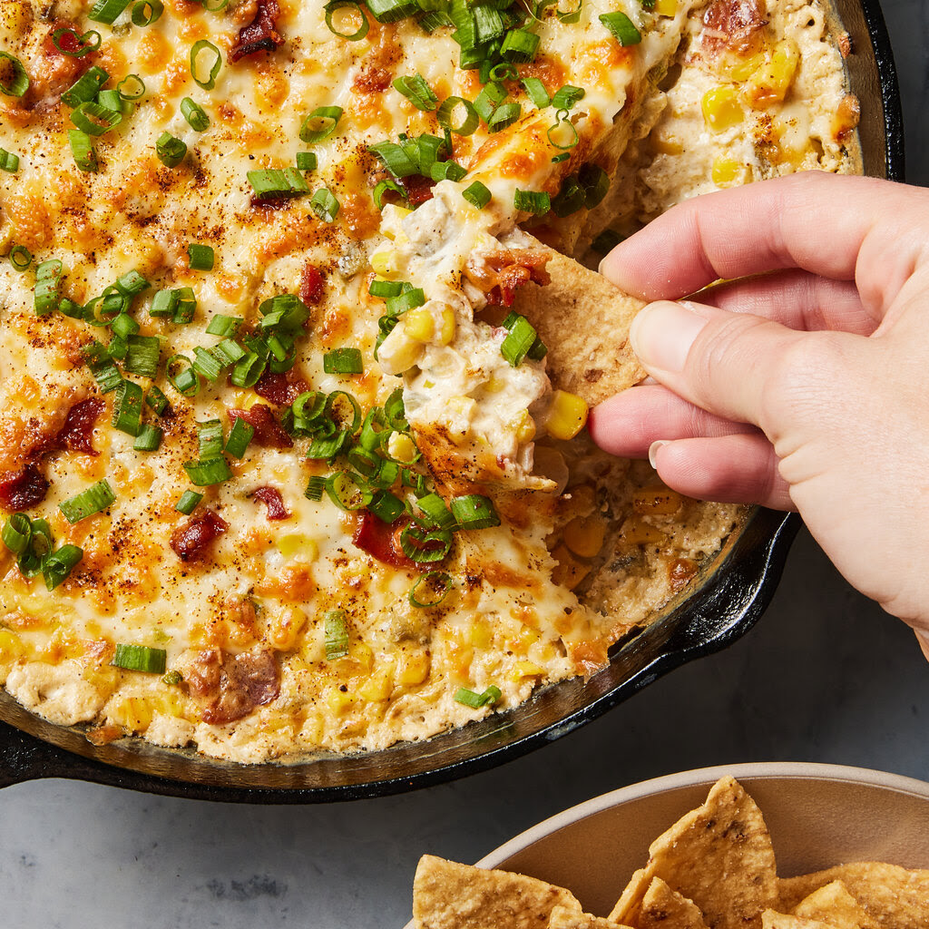 A hand plunges a tortilla chip into a skilled full of creamy corn dip.